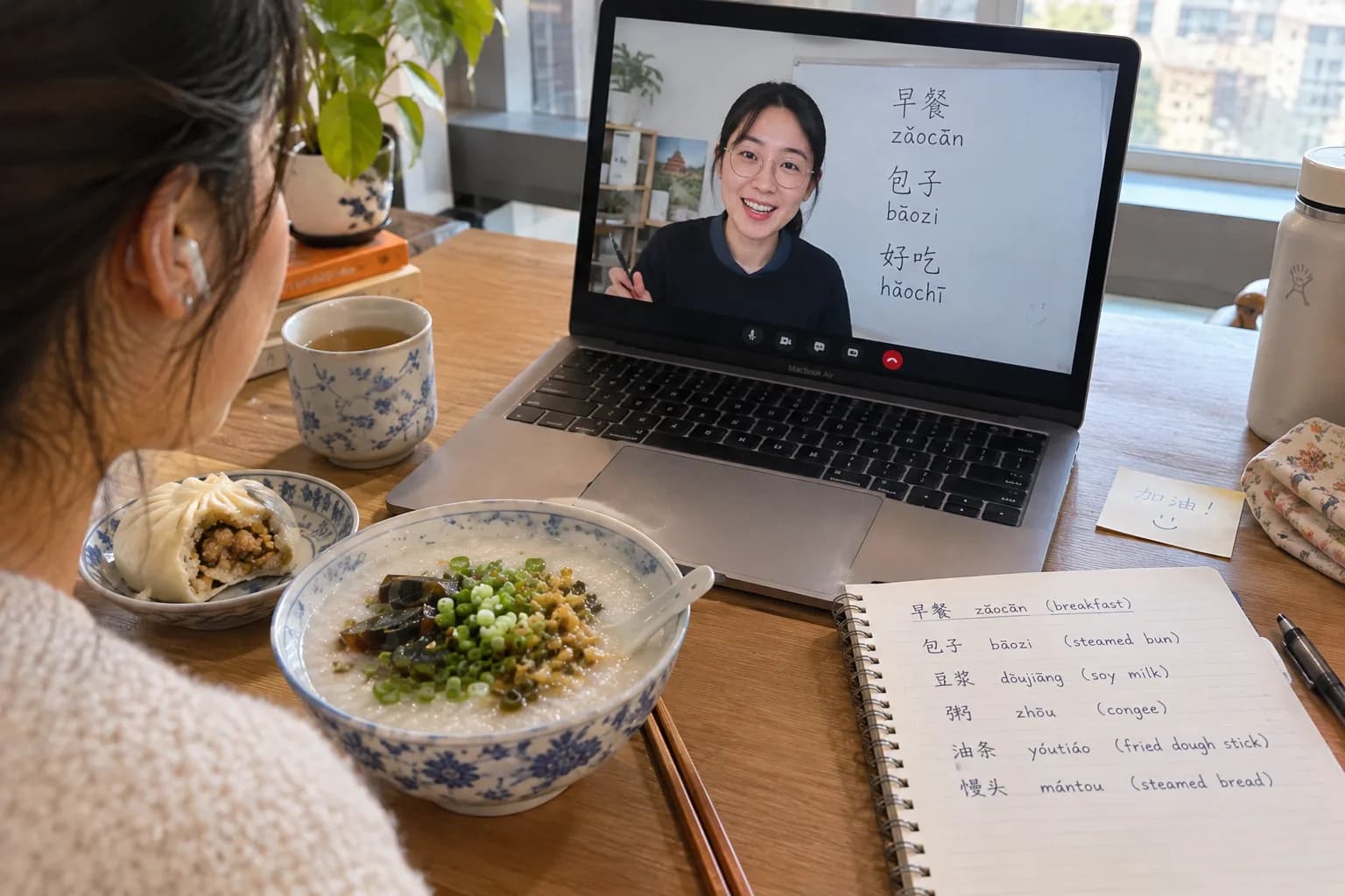 A learner eating congee and dumplings at a desk while a Mandarin teacher on a laptop shows the words 早餐, 包子 and 好吃